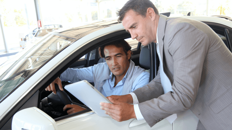 Man sitting in a car talking with a car dealership salesman.