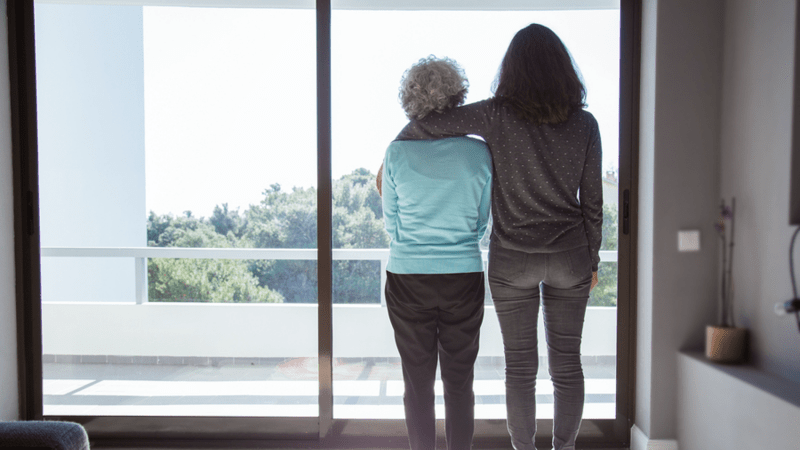 Older woman looking out a window standing next to her adult daughter.