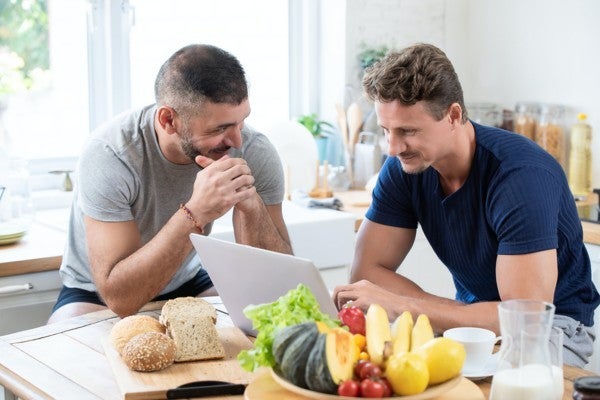 Two men sitting at a table with a laptop and snacks.