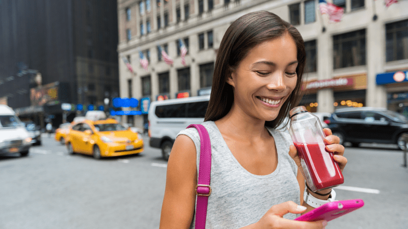 Woman walking down the street looking at her phone and drinking a beverage.