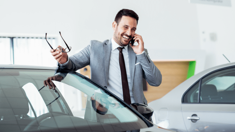 Smiling auto salesman talking on the phone while leaning against a car.